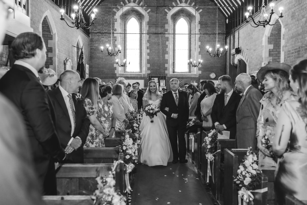 A bride in a white dress walks down the aisle of a decorated church, arm in arm with an older man, as wedding guests seated in pews look on and smile. The scene is captured in black and white.