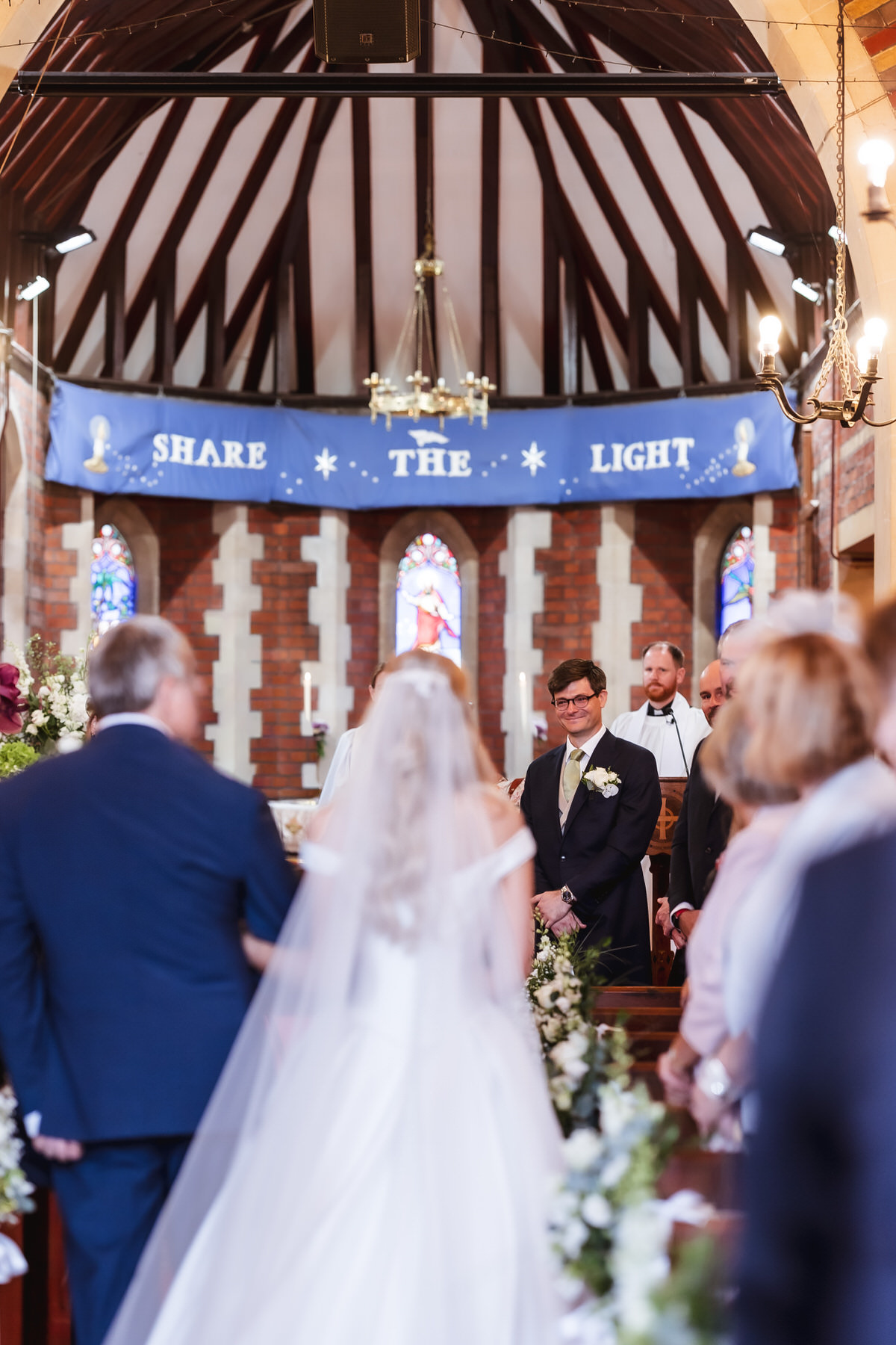 A bride walks down the aisle of a church filled with guests. The groom, smiling, waits at the altar with the officiant. Above them is a blue banner reading “SHARE THE LIGHT.” The church is decorated with flowers.