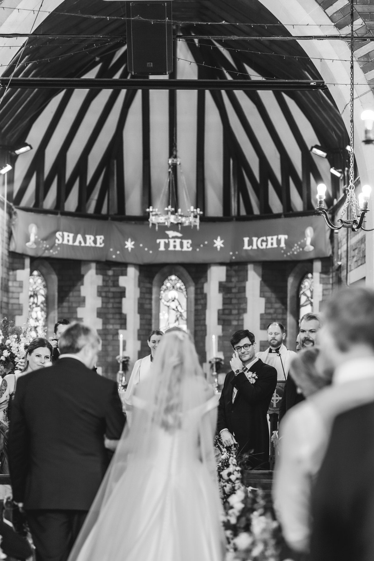 A bride walks down the aisle with a man in a suit at her side in a church, as the groom wipes away tears. Above the altar, a banner reads “SHARE THE LIGHT.” The scene is joyful and emotional.