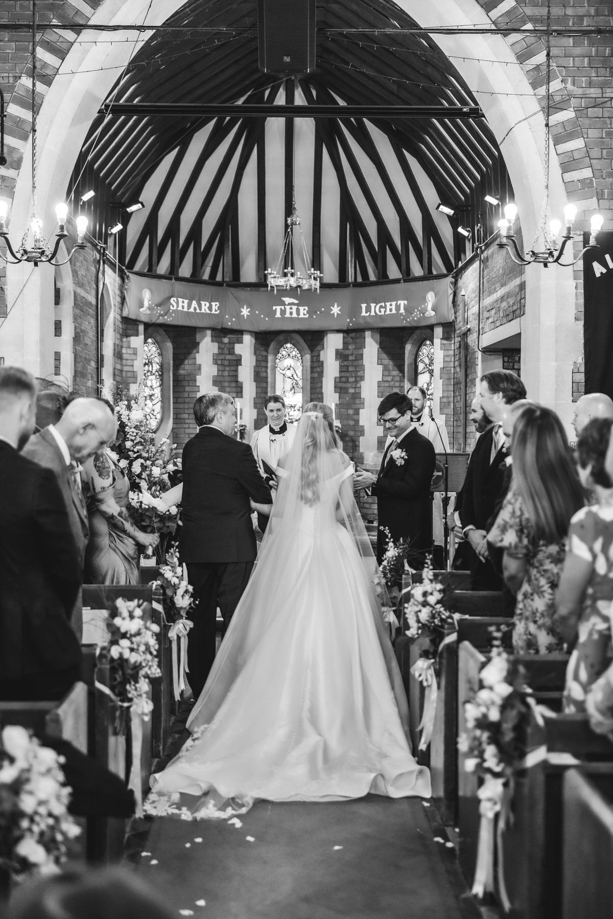 A bride in a wedding gown walks down the aisle of a decorated church, accompanied by an older man. Guests stand on either side, and the groom waits at the altar. The banner reads Share The Light.