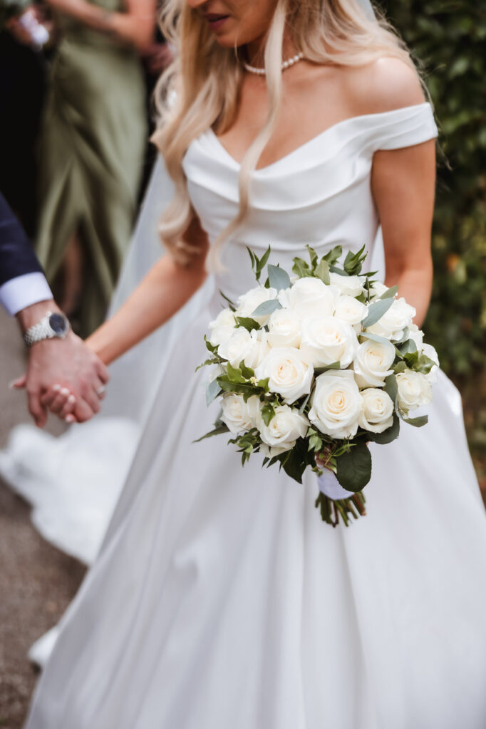 A bride in an off-the-shoulder white dress holds a bouquet of white roses and greenery while holding hands with someone, likely her partner. The background shows another person in a green dress.