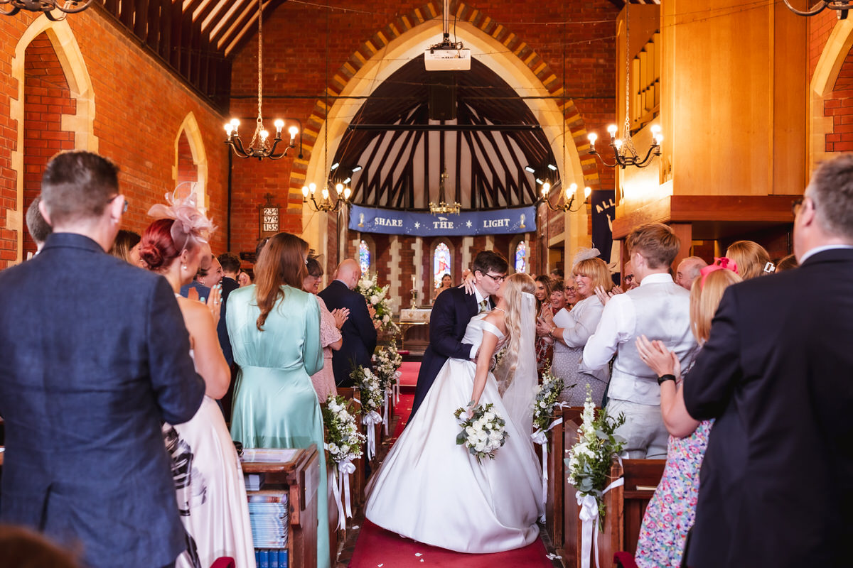 A bride and groom share a kiss at the end of a church aisle while guests stand and applaud, celebrating their wedding ceremony in a decorated, warmly lit church.