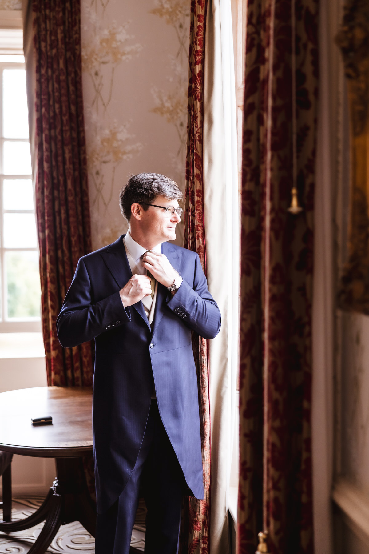 A man wearing a navy suit adjusts his tie while standing by a window with red patterned curtains, looking outside. Soft daylight illuminates the elegant room with a wooden table in the background.