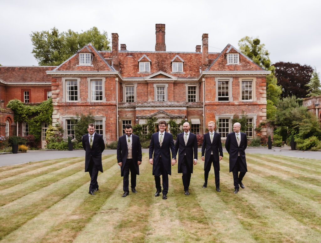 Six men in formal suits walk on a striped lawn in front of a large, old brick mansion with chimneys and tall windows under an overcast sky.
