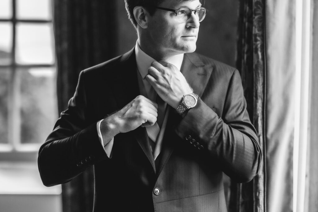 A man in a suit and glasses adjusts his tie while standing near a window. The black and white photo highlights his watch and thoughtful expression as natural light streams in.