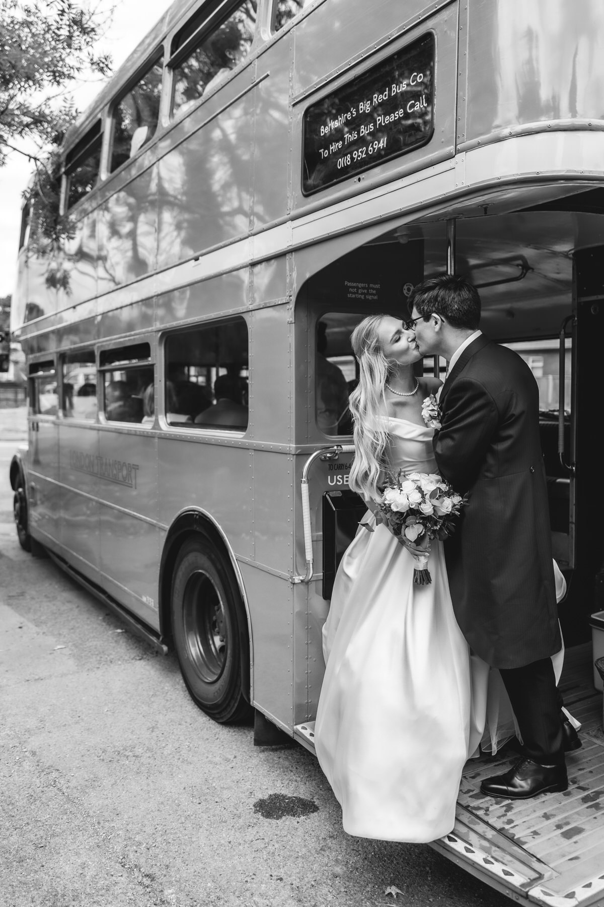 A bride and groom share a kiss at the open door of a vintage double-decker bus. The bride holds a bouquet, and both wear formal wedding attire. The image is in black and white.