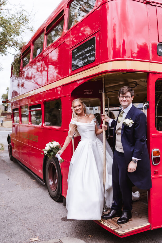 A bride in a white dress and a groom in a suit, both smiling, stand on the open platform at the back of a classic red double-decker bus. The bride holds a bouquet and the groom wears a boutonniere.
