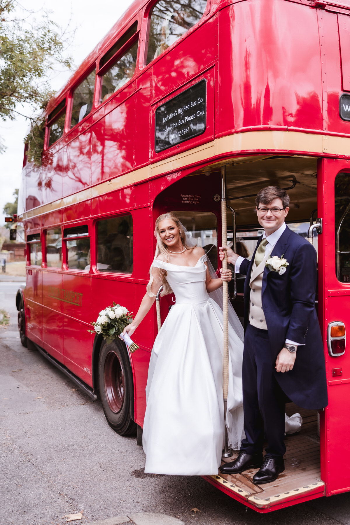A bride in a white dress and a groom in a suit, both smiling, stand on the open platform at the back of a classic red double-decker bus. The bride holds a bouquet and the groom wears a boutonniere.