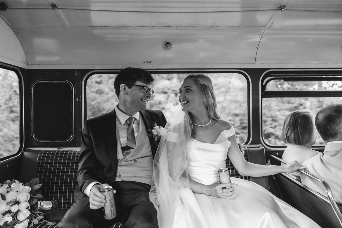 A bride and groom in wedding attire sit smiling at each other on a bus, each holding a drink. The bride wears a white gown and veil, the groom wears a suit. Other passengers are visible in the background.