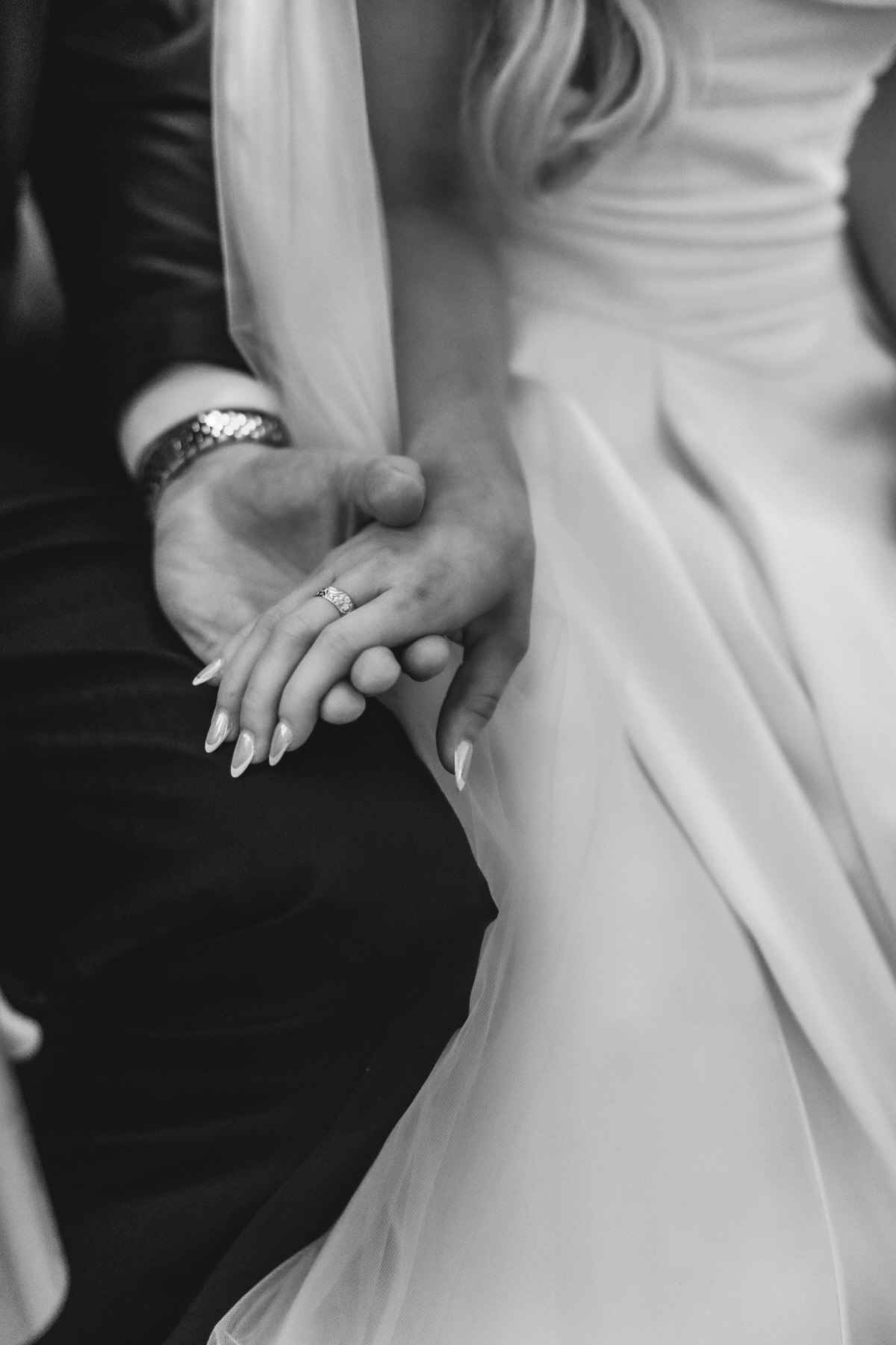 A black and white close-up of a couple holding hands, with the woman wearing a wedding dress and an engagement ring visible on her finger. The man is dressed in a suit.