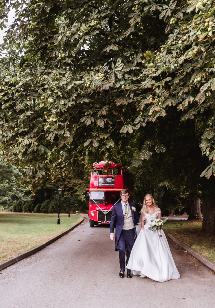 A bride and groom walk arm in arm down a tree-lined path, smiling. A vintage red double-decker bus is parked behind them on the road. Lush green trees arch overhead.