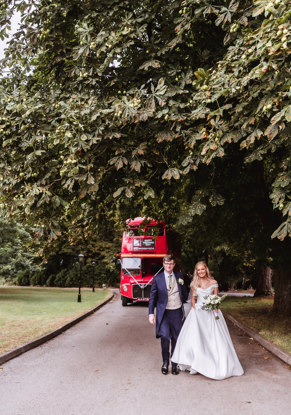 A bride and groom walk arm in arm down a tree-lined path, smiling. A vintage red double-decker bus is parked behind them on the road. Lush green trees arch overhead.
