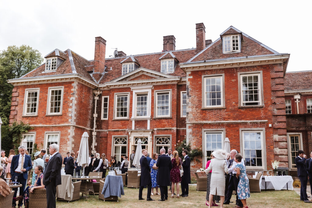 A group of people dressed formally gather and converse in the garden of a large, historic red-brick mansion with many windows and chimneys on a clear day. Tables and chairs are set up on the lawn.