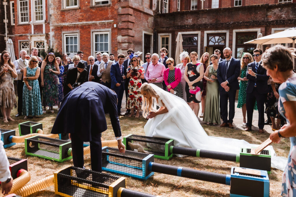A bride and groom participate in an outdoor ferret race game, surrounded by well-dressed wedding guests who watch and smile in a courtyard with brick buildings in the background.