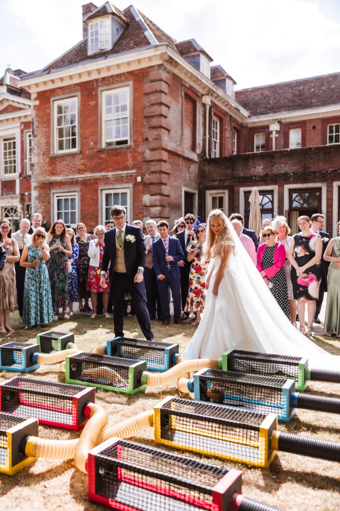A bride and groom play a garden game with colorful boxes and tubes outside a large, historic building, surrounded by a crowd of well-dressed wedding guests watching them.