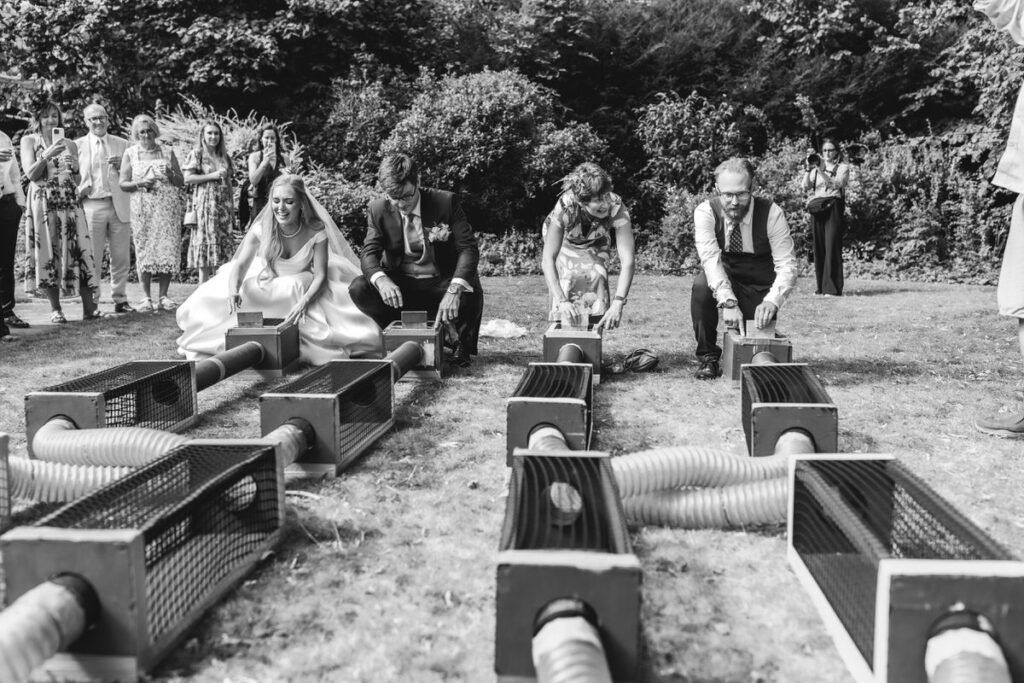 A bride, groom, and two guests crouch on the grass, ready to race small animals through tube mazes, while other guests watch and smile at an outdoor wedding event.