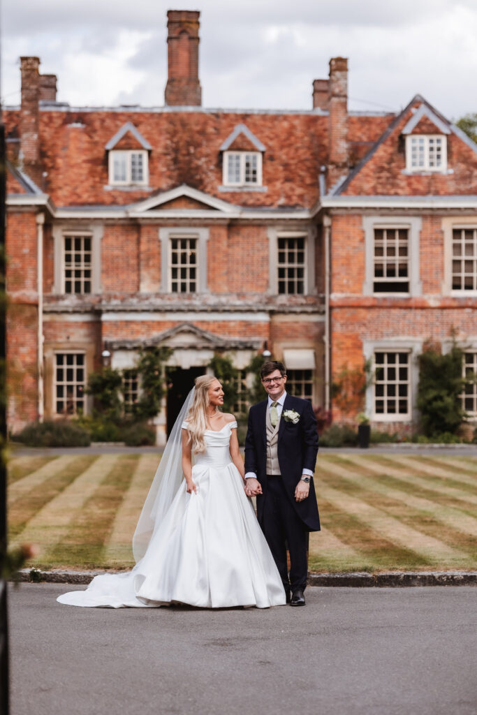 A bride in a white gown and veil stands holding hands with a groom in a dark suit and boutonniere, smiling in front of a large, historic brick mansion with manicured lawns.