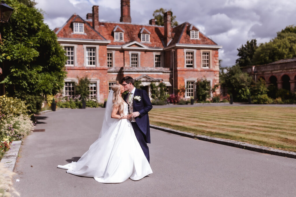 A bride and groom share a kiss outside a large, historic red-brick mansion surrounded by gardens and greenery on a cloudy day. The bride wears a white gown and veil; the groom is in a dark formal suit.