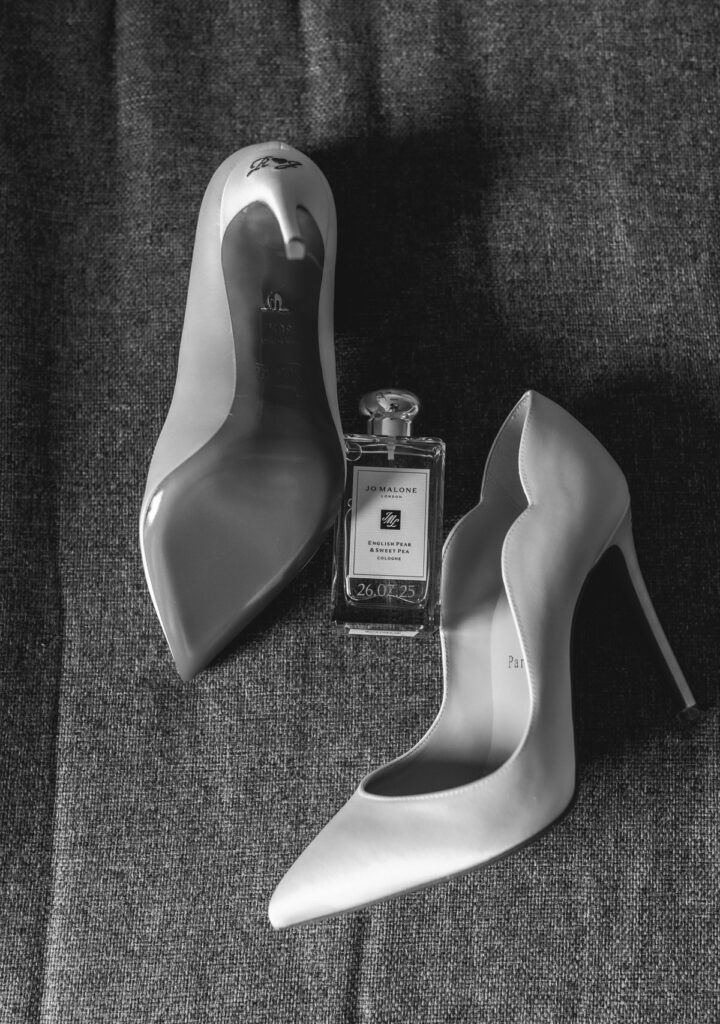 Black and white photo of two high-heeled shoes next to a bottle of Jo Malone perfume, all resting on a textured fabric surface.