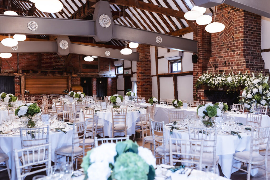 A rustic wedding reception hall with exposed brick and beams, round tables covered in white cloths, white chairs, and elegant green and white floral centerpieces. Soft lighting creates a warm, inviting atmosphere.