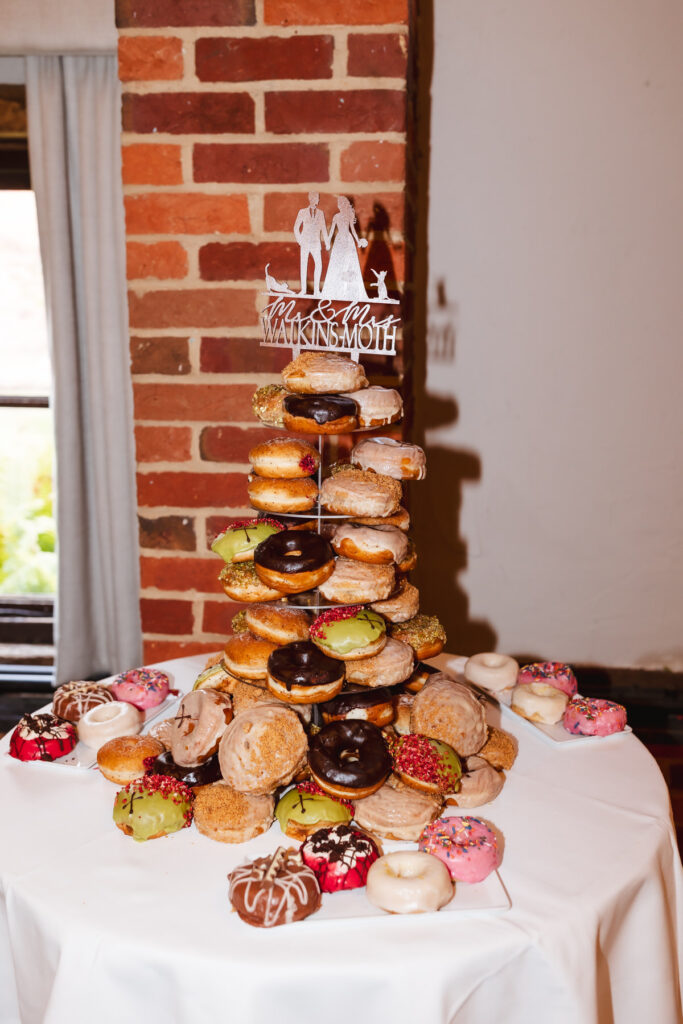 A tiered donut tower with assorted colorful donuts sits on a white tablecloth, topped with a bride and groom cake topper that reads Mr & Mrs Makinson in a room with brick walls and a curtained window.