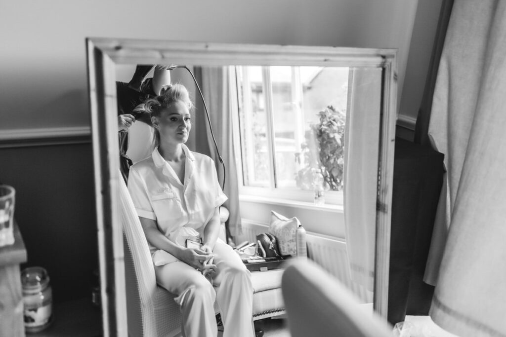 A woman in light-colored clothes sits on a chair, reflected in a large mirror. She looks thoughtful, with curled hair, in a well-lit room with a window and curtains. The image is in black and white.