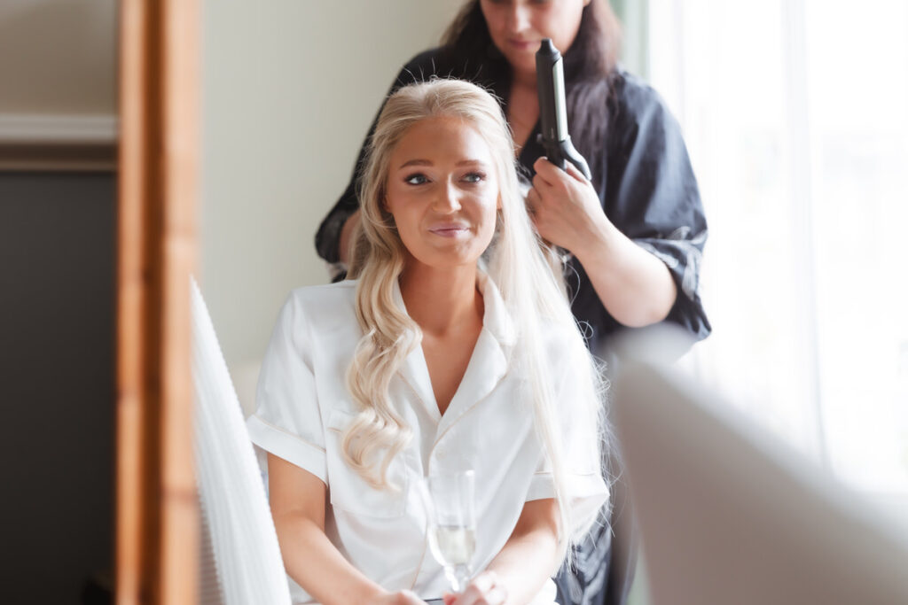 A woman in a white robe with long blonde hair sits smiling, holding a glass, while another woman styles her hair with a curling iron near a window with natural light.