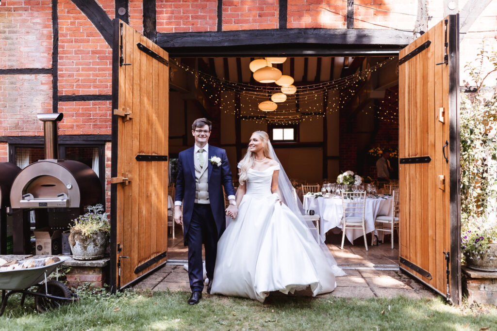 A bride and groom, smiling and holding hands, stand outside an open barn decorated with string lights and set tables, suggesting a rustic wedding reception inside.