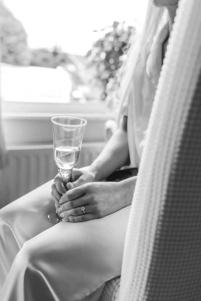 A person in a light-colored outfit sits by a window, holding a champagne flute with both hands; an engagement ring is visible on their left hand. The image is in black and white.
