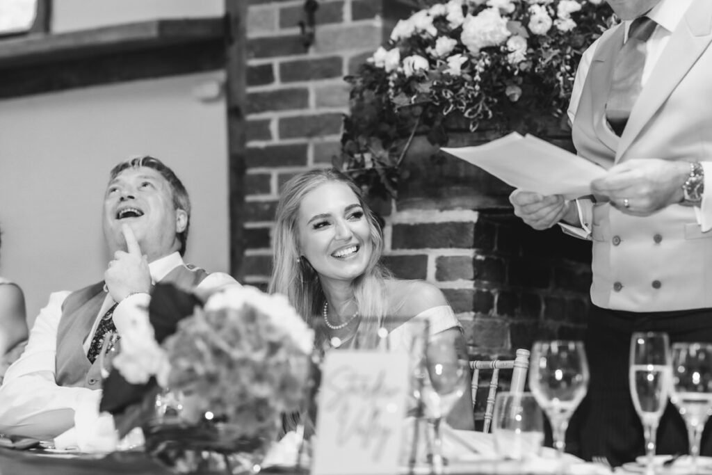 A black-and-white photo of a wedding reception shows a smiling woman sitting between a laughing man and a person standing and reading from a paper. Flowers and glasses decorate the table in front of them.