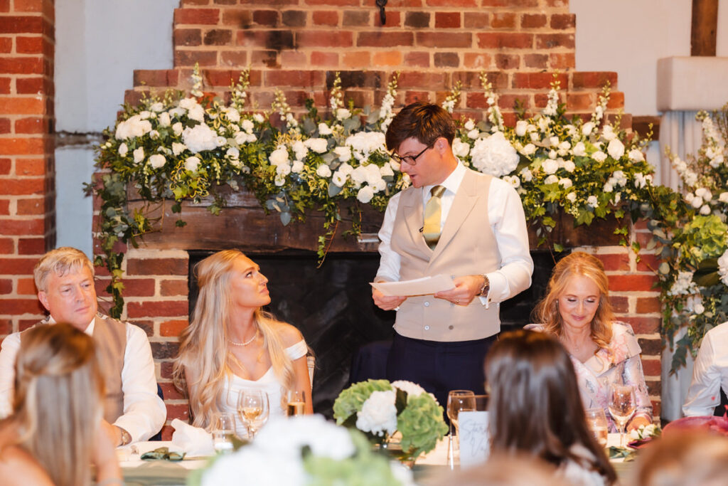 A man in a vest and tie stands holding a speech in front of a seated wedding party at a decorated table, with white flowers and greenery on a brick fireplace backdrop. The guests look at him, smiling and attentive.