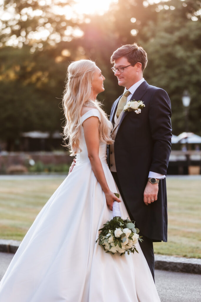 A bride in a white gown and a groom in a dark suit smile at each other outdoors, with the bride holding a bouquet of white flowers and greenery; sunlight filters through the trees in the background.