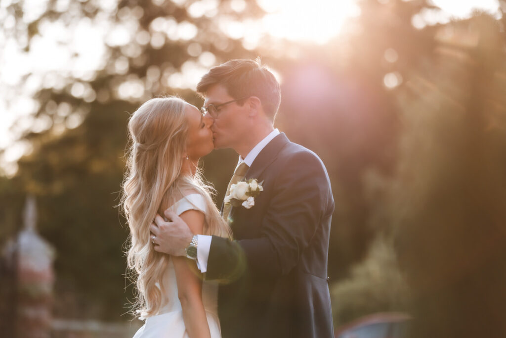A bride and groom share a kiss outdoors at sunset, surrounded by soft golden light. The groom wears a dark suit with a boutonnière, while the bride has long wavy hair and a white dress.