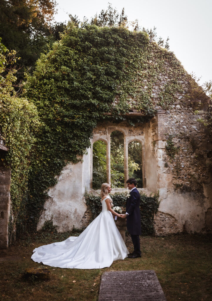 A bride and groom stand facing each other, holding hands, in front of an ivy-covered stone wall with tall arched windows, outdoors, surrounded by greenery.