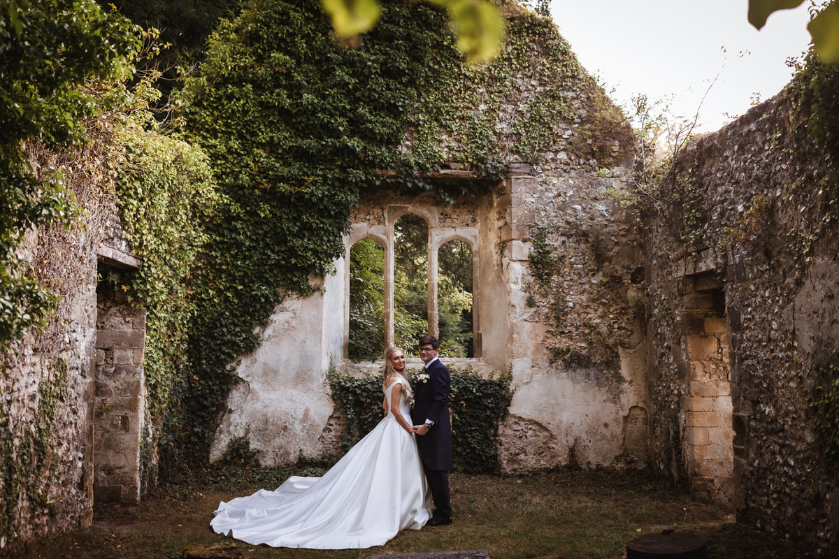 A bride and groom stand holding hands in front of an old, ivy-covered stone wall with arched windows, surrounded by lush greenery and the ruins of a historic building.