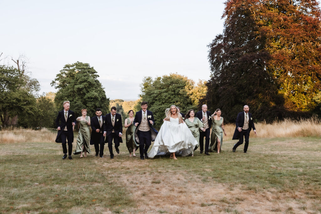 A bride in a white dress and a groom in a tuxedo walk on grass with their wedding party, all smiling and dressed in formal attire, surrounded by trees and a natural outdoor setting.
