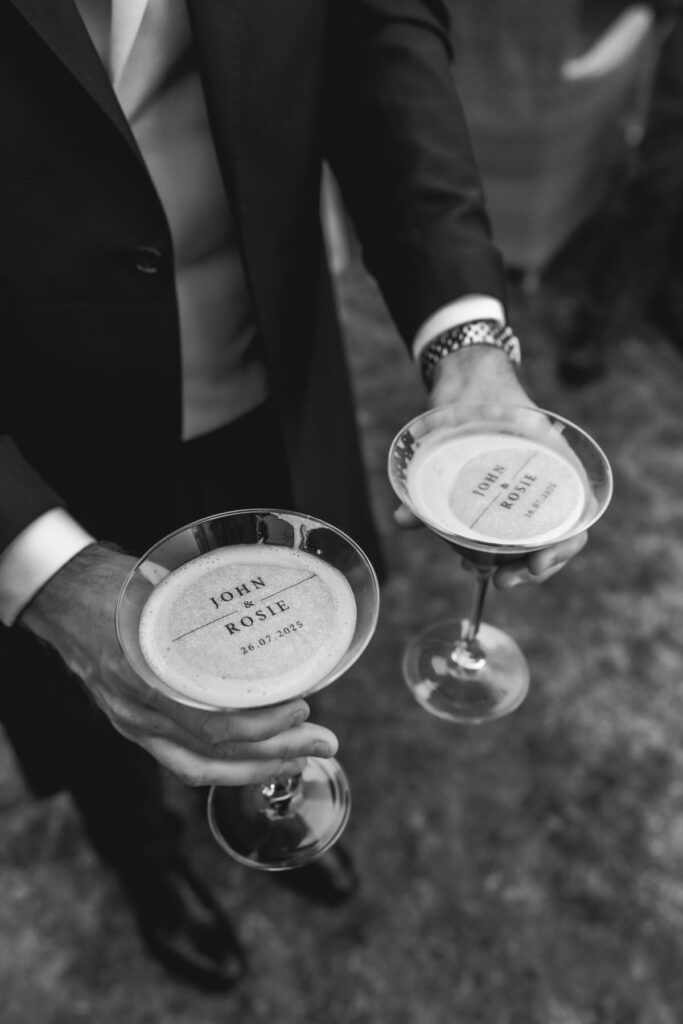 A person in a suit holds two champagne glasses with frothy tops. The foam on each drink is printed with the names John & Rosie and a date, 24-07-2019. The photo is in black and white.