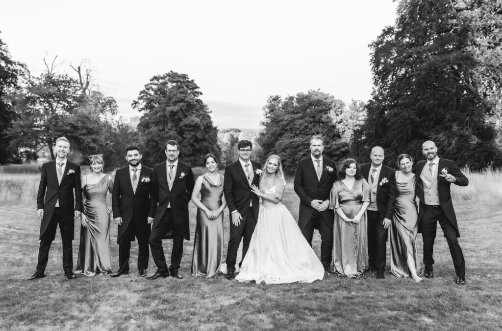 A black-and-white photo of a wedding party posing outdoors. The group, dressed in formal attire, stands in a row on grass with trees in the background. The bride and groom are centered, surrounded by bridesmaids and groomsmen.