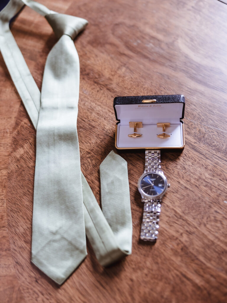 A light green necktie, a silver wristwatch, and a boxed set of gold cufflinks are arranged on a wooden surface.