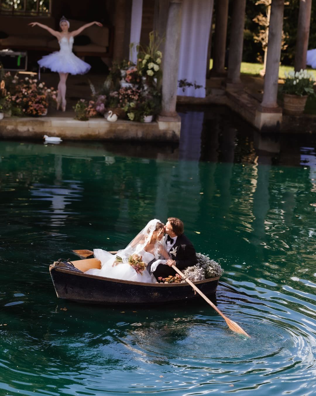 A newlywed couple kisses in a small rowboat adorned with flowers on a turquoise pond, captured by a Hampshire wedding photographer. In the background, a ballerina in a white tutu poses gracefully on the shore near columned architecture.