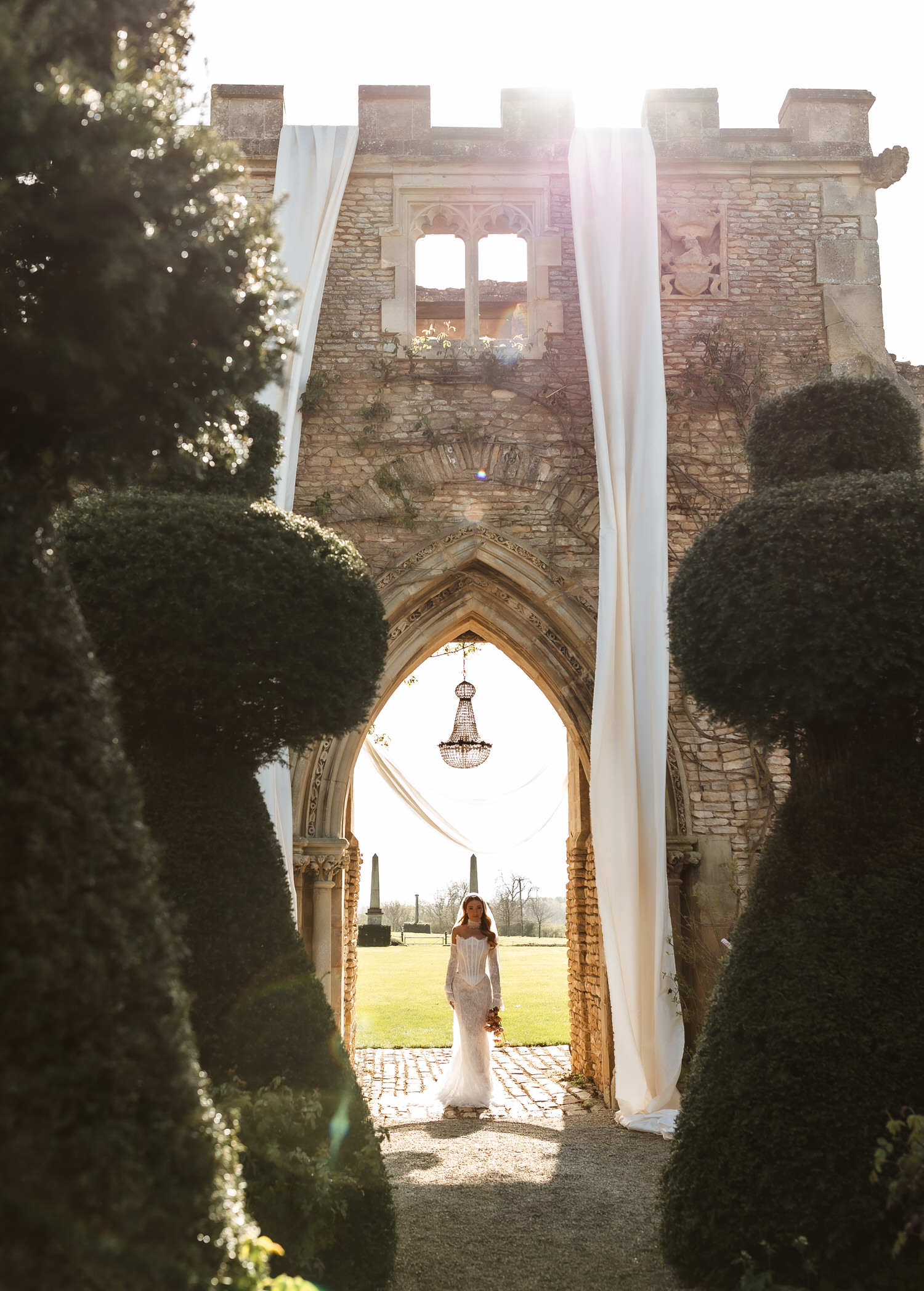 A bride in a long white gown stands under a stone archway adorned with flowing white drapes and a chandelier, framed by manicured topiary trees and sunlight—an elegant scene perfect for a country-house wedding photographer.