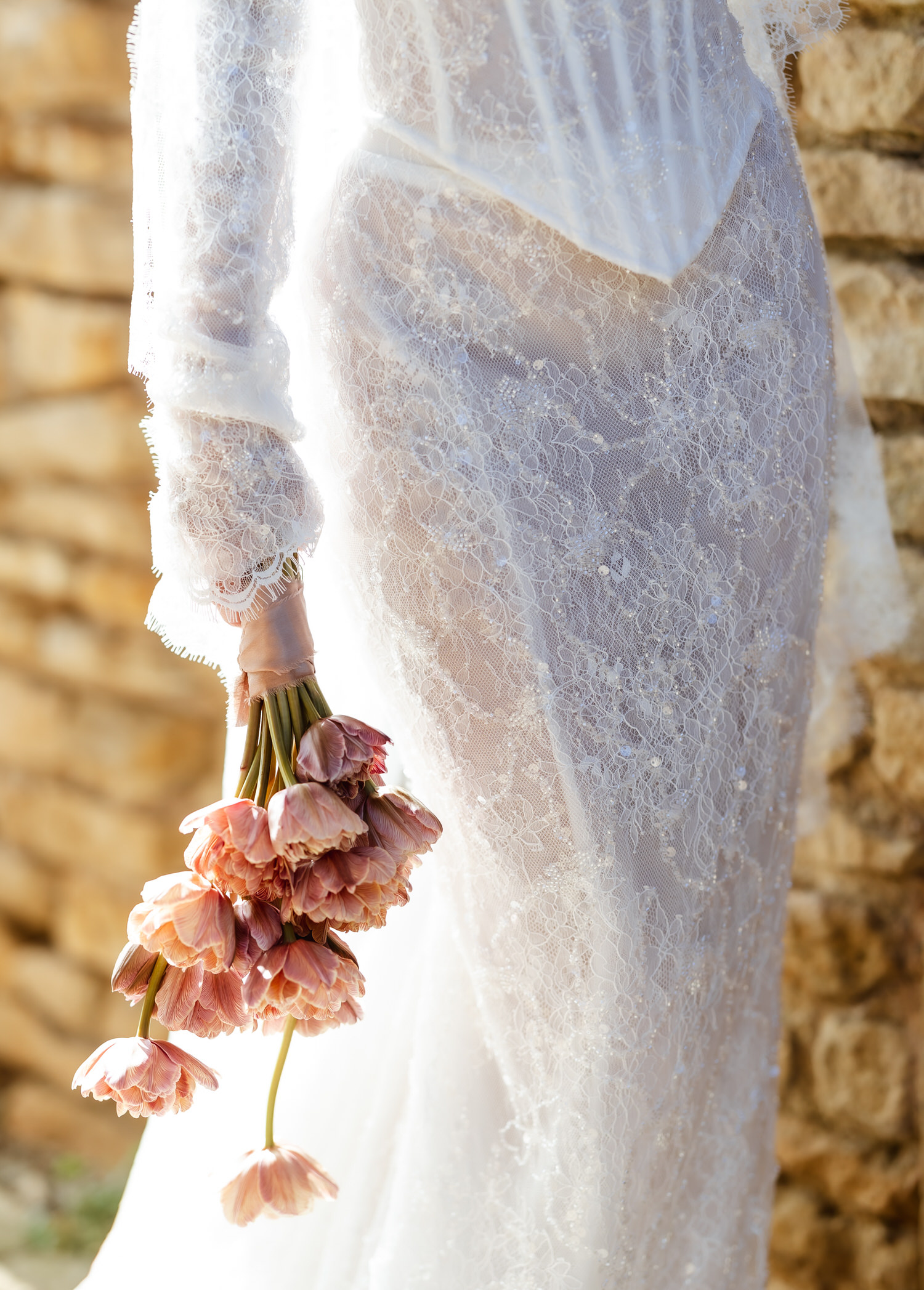 A person in a sheer, lace wedding dress holds a bouquet of pink drooping tulips, standing beside a rustic stone wall in soft sunlight—perfect for any country-house wedding photographer to capture.
