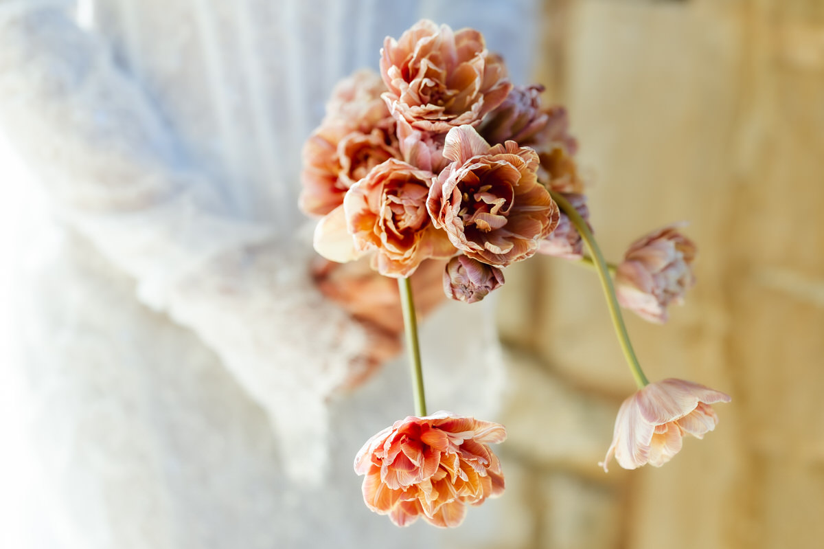 Close-up of a person in a white lace dress holding a bouquet of pink and peach double tulips, captured by a Hampshire wedding photographer against a softly blurred, warm-toned background.
