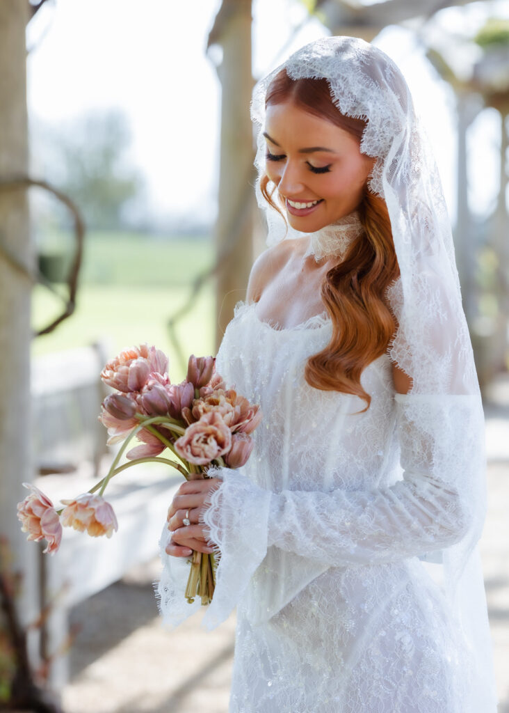 A bride in a long-sleeved lace wedding dress and veil smiles while holding a bouquet of light pink flowers outdoors on a sunny day, beautifully captured by a Hampshire wedding photographer.