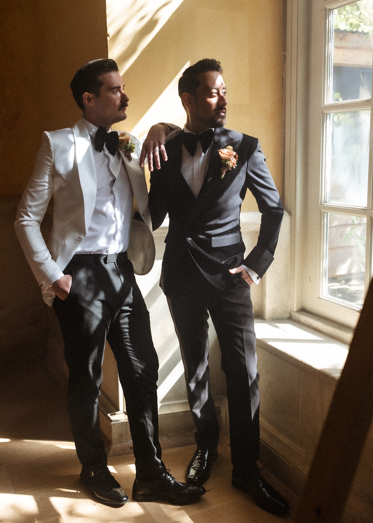 Two men in tuxedos, one in a white jacket and the other in black, stand by a sunlit window. Captured by a Hampshire wedding photographer, they pose confidently with relaxed expressions and hands in their pockets.