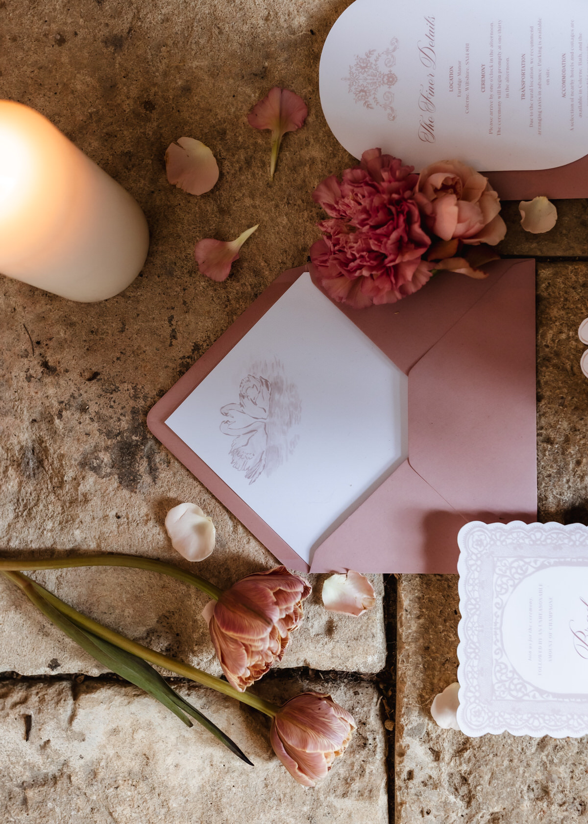 A pink envelope with a swan illustration, floral wedding invitations, pink flowers, rose petals, and a lit candle are beautifully arranged on a stone surface—perfect inspiration for any Hampshire wedding photographer.