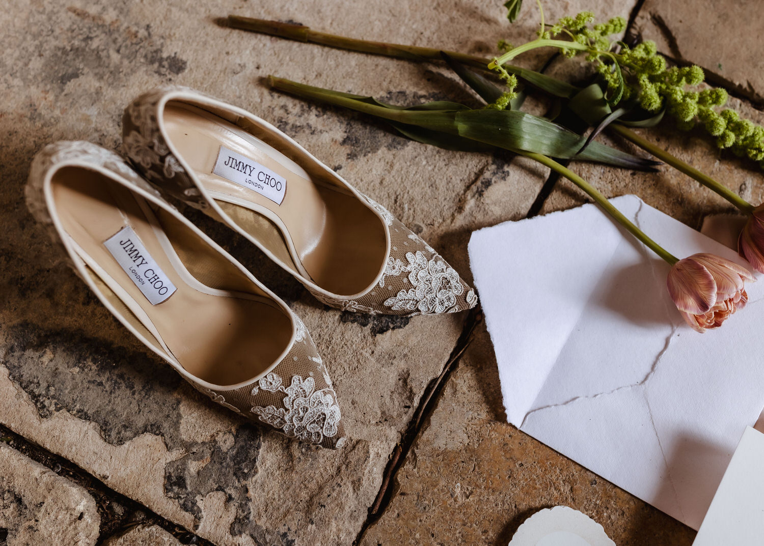 A pair of lace-patterned Jimmy Choo shoes rest on a stone floor beside a white envelope, pink tulips, and green foliage—an elegant detail often captured by a country-house wedding photographer.