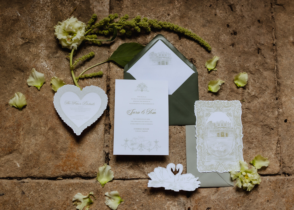 A flat lay of elegant wedding invitations, envelopes, and white paper swans, arranged on a rustic stone surface with scattered green flowers and petals—perfect inspiration for a Hampshire wedding photographer capturing timeless details.