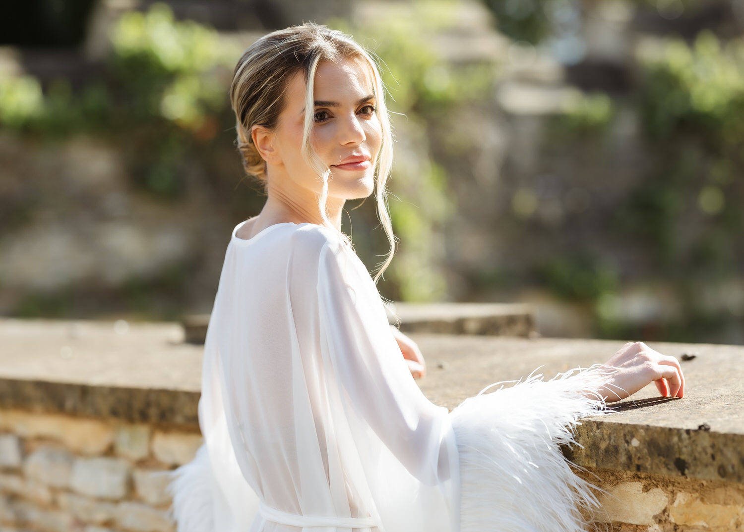 A woman in a white, feather-trimmed dress stands outdoors by a stone wall, looking over her shoulder at the camera. Sunlight softly illuminates her face as a country-house wedding photographer captures the blurred greenery in the background.