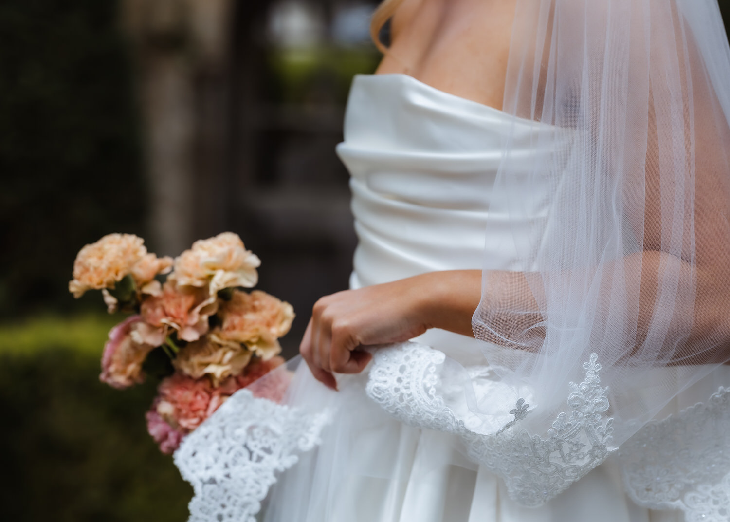 A bride in a strapless white wedding dress and veil holds a bouquet of light pink flowers, standing outdoors with greenery—a perfect moment captured by a country-house wedding photographer.